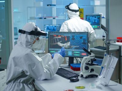 Chemist doctor in ppe suit working at pc while lab technician bringing her blood samples. Team of scientists examining vaccine evolution with high tech for researching treatment against covid19 virus