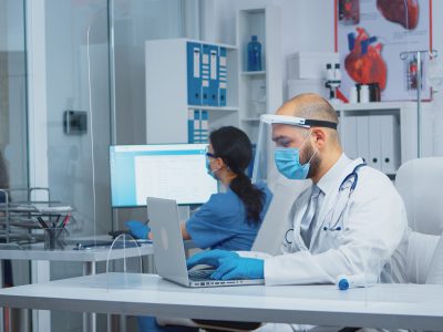Doctor with protection mask and visor typing the treatment on laptop while mother comes with daughter to the consultation in hospital during coronavirus pandemic. Equipped nurse talking with patients.